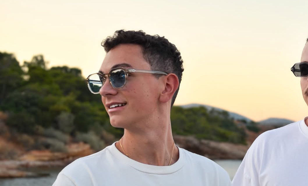 Two men wearing white t-shirts and sunglasses standing outdoors near the water at sunset. The man in the foreground wears a white crewneck t-shirt with a small black graphic on the left chest and silver-framed sunglasses.