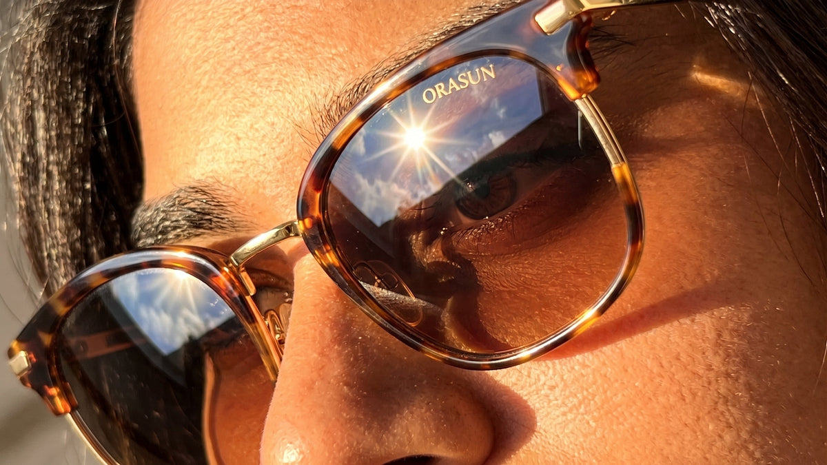 Close-up of a person wearing tortoiseshell sunglasses with gold metal frames and dark lenses, with the brand name ORASUN visible on the lens.